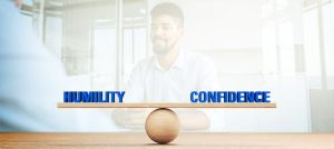 the words Humility and Confidence are balanced over a wooden sphere scale