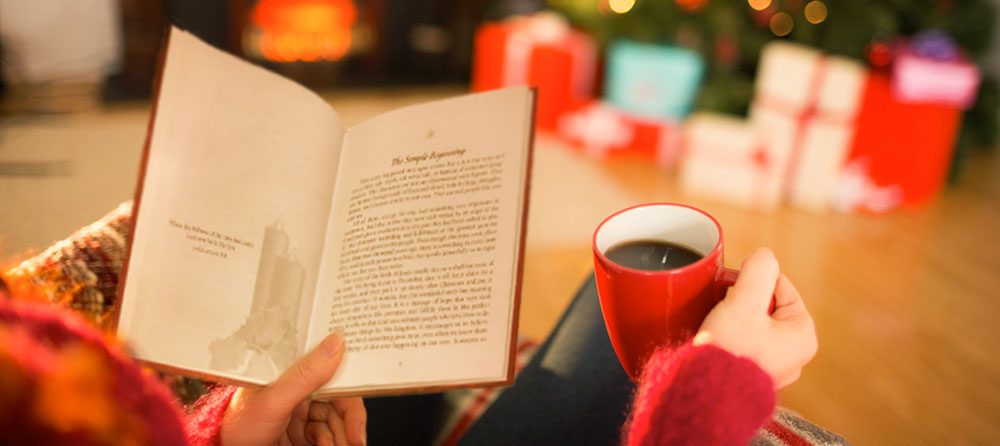 woman holding coffee reading book in front of Christmas Tree