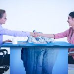two women sitting at a desk shaking hands with an image of an iceberg overlaid on the desk