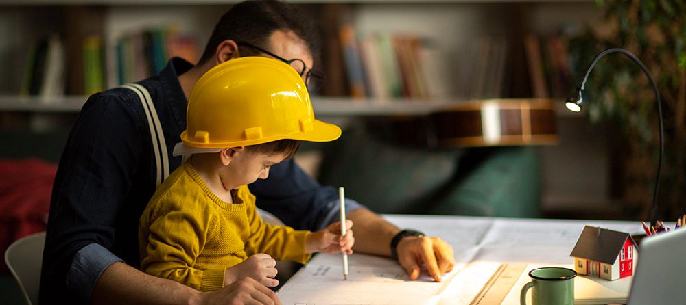 construction pro sitting on desk with son wearing a hardhat sitting on his lap